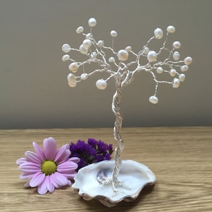 May include: A silver wire tree with white pearl beads, standing on a white seashell base. The tree is on a wooden surface with a pink flower and purple flowers in the foreground.