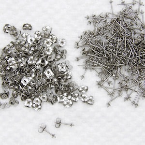 Op de afbeelding: Een grote verzameling zilverkleurige metalen oorbelonderdelen, inclusief een hoop vlindervormige oorbelachterkantjes en een aparte hoop oorbelstiften met platte schijfjes, plus twee volledig gemonteerde oorknopjes onderaan.