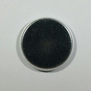 May include: A round, silver-colored button with a brushed metal surface. The button has a slightly raised, textured edge and a dark gray center. The button is set against a white background.