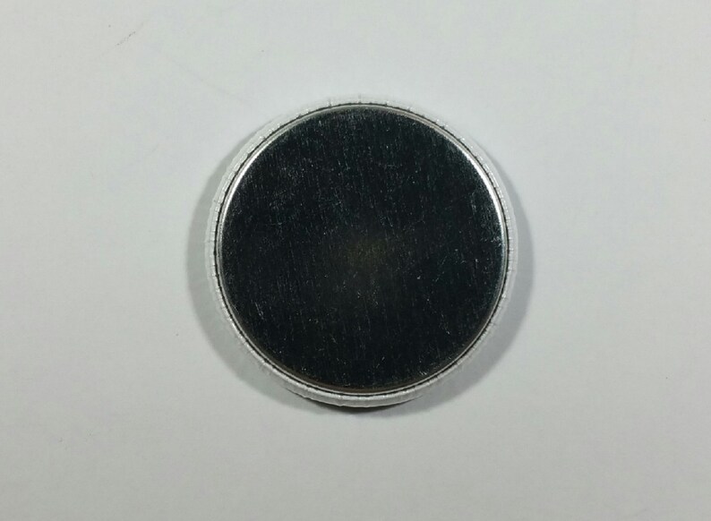 May include: A round, silver-colored button with a brushed metal surface. The button has a slightly raised, textured edge and a dark gray center. The button is set against a white background.