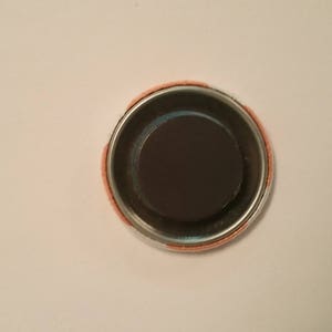 May include: A round magnet with a dark brown center and a silver metal rim. The outer edge has a peach-colored fabric. The magnet is designed to attach to metal surfaces.