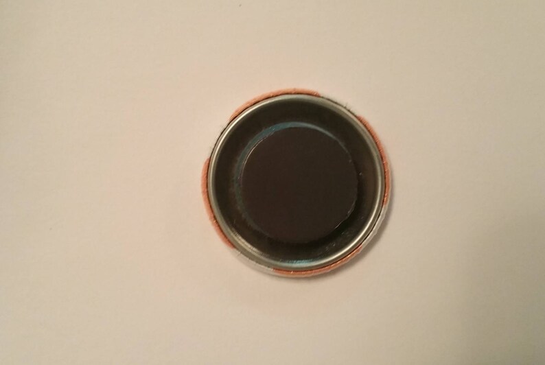 May include: A round magnet with a dark brown center and a silver metal rim. The outer edge has a peach-colored fabric. The magnet is designed to attach to metal surfaces.