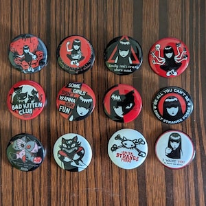 May include: Twelve black and white button pins with red accents featuring a cartoon character with black hair and a black dress. The pins have various sayings including "Bad Kitten Club", "Some Girls Don't Wanna Have Fun", "Emily Isn't Crazy She's Mad", and "Be All You Can't Be Emily Strange Posse".