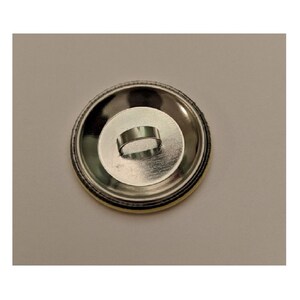 May include: A round, silver-colored button back with a metal loop for attachment. The button has a circular design with a yellow and black edge.