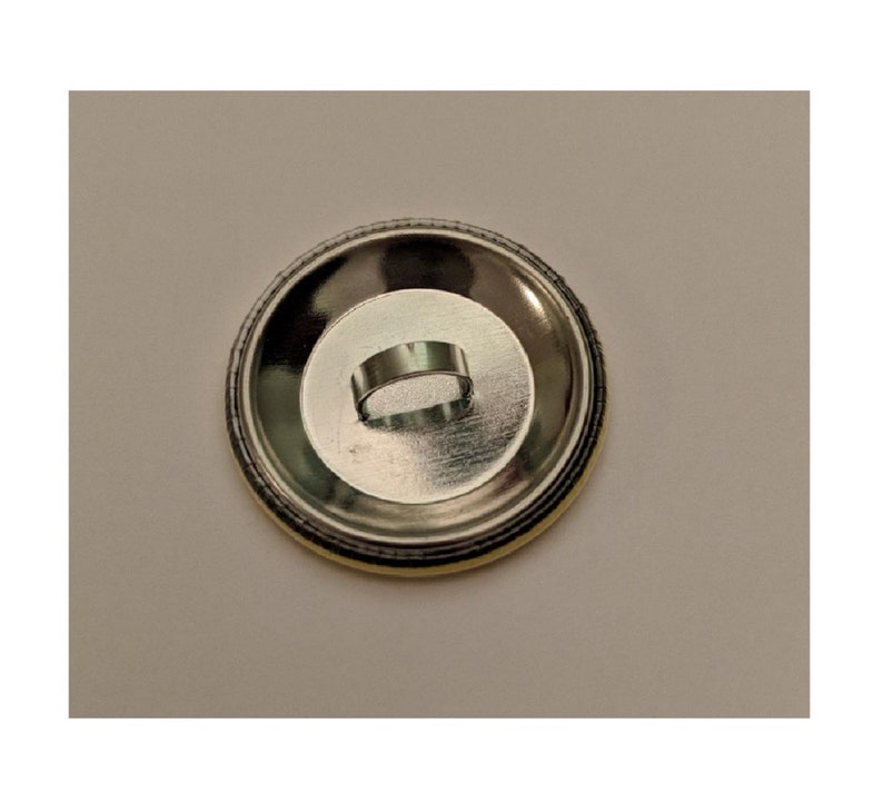 May include: A round, silver-colored button back with a metal loop for attachment. The button has a circular design with a yellow and black edge.