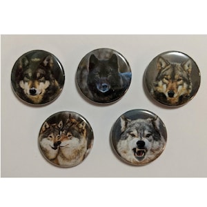 May include: Set of five round buttons featuring wolf portraits. The buttons display various wolf faces, including a black wolf and wolves with brown and gray fur. The buttons are on a white background.