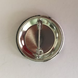 May include: A round button pin with a silver metal back and a safety pin clasp. The button's edge is gray, and the back is shiny silver. The button is on a white surface.