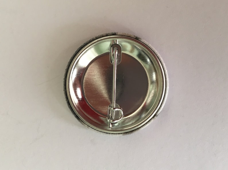 May include: A round button pin with a silver metal back and a safety pin clasp. The button's edge is gray, and the back is shiny silver. The button is on a white surface.
