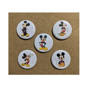 May include: Five white round buttons featuring different poses of Mickey Mouse. Mickey is depicted in various outfits and poses, with his signature black ears, red shorts, and yellow shoes. The buttons are arranged on a brown surface.