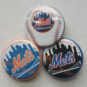 May include: Three round Mets baseball team buttons. One button is white with red stitching and the Mets logo. The other two buttons are blue and orange with the Mets logo and a skyline.