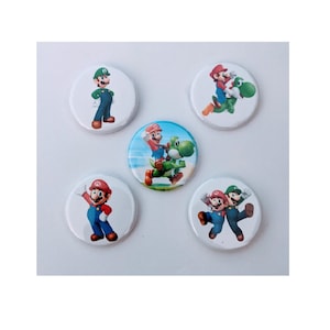 May include: A collection of five round buttons featuring colorful illustrations of characters from the Super Mario video game series. The buttons showcase Mario, Luigi, and Yoshi in various action poses, set against white backgrounds.
