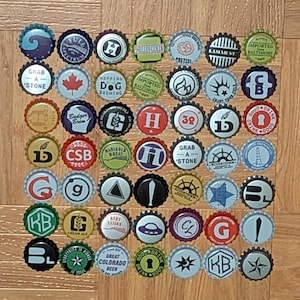 May include: A collection of forty-two assorted metal bottle caps arranged in a grid. Each cap features unique graphic designs, including various logos, text, symbols, and illustrations in diverse colors such as red, blue, green, black, and white.