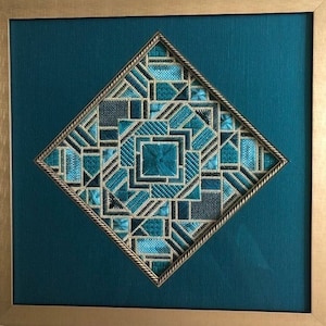 Op de afbeelding: Een ingelijst, ruitvormig stuk naaldkunst met een geometrisch patroon in tinten teal en goud.