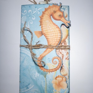 May include: A handmade card with a blue background and a seahorse illustration. The seahorse is orange and brown with a white belly. The card is tied with twine and has a white tag.