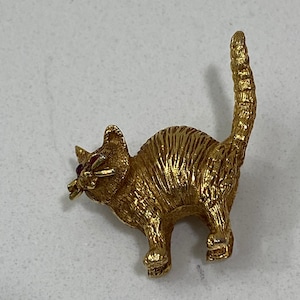 May include: A gold-toned cat figurine with red eyes. The cat is standing on all fours with its tail curled up.