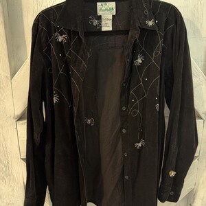 May include: A black long-sleeve button-down shirt featuring embroidered silver spider web patterns and small spider motifs across the front. The shirt has a classic collar, a front button placket, and a label at the neck with three small bird icons.