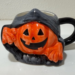 May include: A ceramic mug shaped like a jack-o'-lantern wearing a black hooded cloak. The pumpkin face is bright orange with black triangular eyes and a wide, toothy grin. The mug features a black handle and a dark, textured hood design.