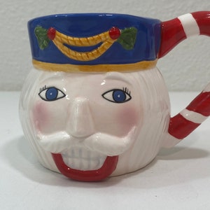 May include: A white ceramic mug shaped like a nutcracker's face. The mug has a blue hat with a gold trim and a red and white candy cane handle.