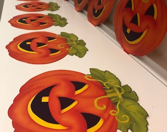 May include: A set of nine orange paper pumpkins with black eyes, a wide smile, and a yellow outline. Each pumpkin has a green stem with a leaf. The pumpkins are arranged in a row on a white surface.