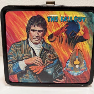 May include: A rectangular metal lunch box featuring a graphic of The Fall Guy with a man in a jacket and a stunt person in a blue helmet against a background of orange flames. The text reads The Fall Guy and Fall Guy Stuntman Assoc.
