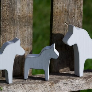 May include: Three white wooden Dala horses, a traditional Swedish folk art, are arranged in a row on a wooden fence. The horses are painted white and have a simple, stylized design.