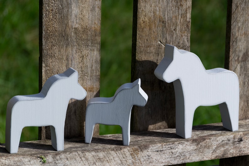 May include: Three white wooden Dala horses, a traditional Swedish folk art, are arranged in a row on a wooden fence. The horses are painted white and have a simple, stylized design.