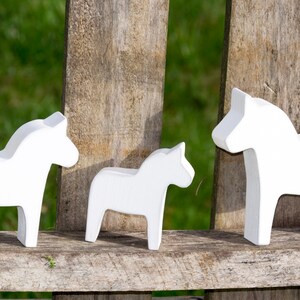 May include: Three white wooden Dala horses, a traditional Swedish folk art, are displayed on a wooden surface. The horses are arranged in a row, with the largest horse on the right, a medium-sized horse in the middle, and the smallest horse on the left.