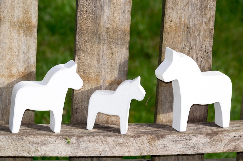 May include: Three white wooden Dala horses, a traditional Swedish folk art, are displayed on a wooden surface. The horses are arranged in a row, with the largest horse on the right, a medium-sized horse in the middle, and the smallest horse on the left.