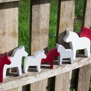 May include: A row of wooden horse figurines, painted in alternating red and white, are displayed on a wooden fence. The horses are all facing the same direction and are arranged in a line.