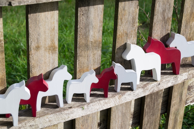 May include: A row of wooden horse figurines, painted in alternating red and white, are displayed on a wooden fence. The horses are all facing the same direction and are arranged in a line.