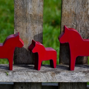 May include: Three red wooden Dala horses, a traditional Swedish folk art, are arranged in a row on a wooden surface. The horses are different sizes, with the largest horse on the right and the smallest horse on the left.