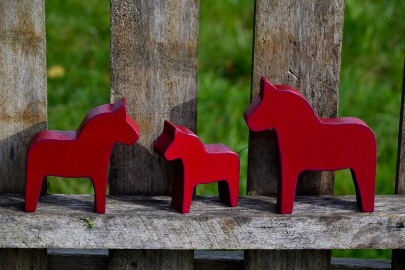 May include: Three red wooden Dala horses, a traditional Swedish folk art, are arranged in a row on a wooden surface. The horses are different sizes, with the largest horse on the right and the smallest horse on the left.
