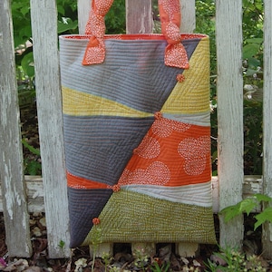 May include: A patchwork tote bag made from orange, grey, yellow and green fabric. The bag has a geometric pattern and is accented with buttons.
