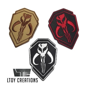 Könnte beinhalten: Drei gestickte Aufnäher mit einem stilisierten Schädeldesign. Einer ist beige mit braunen Details, ein anderer ist schwarz mit weißen Details und der dritte ist schwarz mit roten Details. Die Aufnäher sind schildförmig.