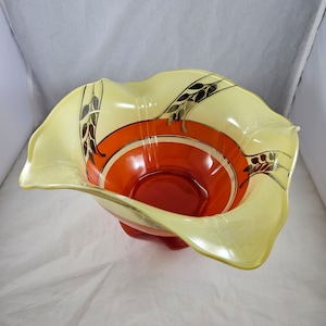 Puede incluir: Un tazón grande de vidrio amarillo con un borde rizado y un interior rojo. El tazón tiene un diseño de tallos de trigo negro y dorado alrededor del borde.