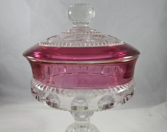 Puede incluir: Un platito de cristal transparente para dulces con una tapa de cristal rosa. El plato tiene una base de pedestal y un borde festoneado. La tapa tiene un pomo de cristal transparente.