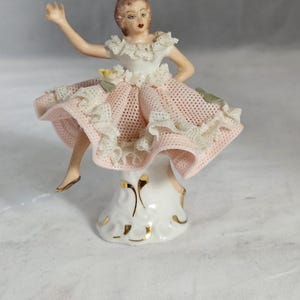 May include: A porcelain figurine of a woman in a pink and white dress, with a ruffled skirt and bodice. The figurine has a delicate design, with gold accents on the base and a detailed face. The woman is posed with one arm raised.