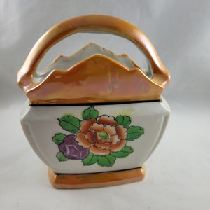 May include: A white ceramic vase with a gold handle and floral design. The vase has a square base and a rounded top. The floral design features a large orange flower with green leaves and a smaller purple flower.