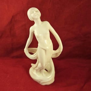 May include: A cream-colored ceramic figurine of a woman with flowing fabric. The figure is in a graceful pose, with arms outstretched and head tilted back. The figurine is on a red background.