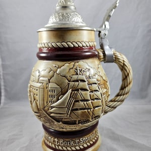 May include: A ceramic beer stein with a detailed relief of a sailing ship and a cityscape. The stein has a pewter lid with a hinged handle and a rope-like handle. The base of the stein reads "antine Bark "T. Staysail".