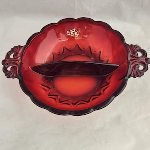 Può includere: Un piatto da portata diviso in vetro rosso rubino con bordo smerlato e manici decorati. Il piatto ha un divisore scuro e un adesivo Viking Hand Made. Il piatto è probabilmente vintage e adatto per servire snack o antipasti.