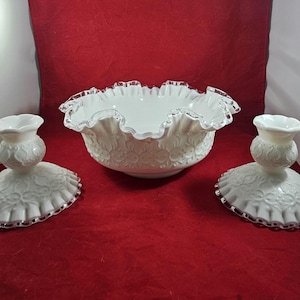 May include: A white, vintage-style centerpiece set with a ruffled-edge bowl and two matching candle holders. The set has a floral pattern and silver trim, creating an elegant aesthetic. The items are displayed on a red background.