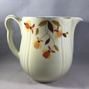 Può includere: Un'anfora in ceramica bianca con un motivo floreale in tonalità arancio, giallo e marrone. L'anfora ha un corpo scanalato e un bordo dorato.