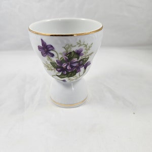 May include: White porcelain egg cup with a gold rim and base. The cup is decorated with a floral design of purple violets and green leaves. The egg cup has a pedestal base and is designed to hold a single egg.