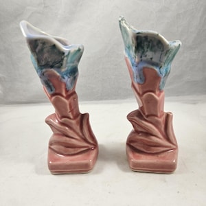 Könnte beinhalten: Zwei Keramik-Blumenvasen. Die Vasen sind staubrosa mit einem blattartigen Design an der Basis und am Stiel. Die Oberteile sind eine Mischung aus Blau, Grün und Weiß. Die Vasen haben eine einzigartige, organische Form.