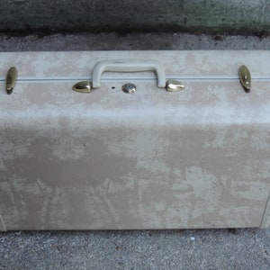 May include: Vintage beige suitcase with a speckled pattern, a cream-colored handle, and gold-toned metal accents. The suitcase has a rectangular shape and is designed for travel.