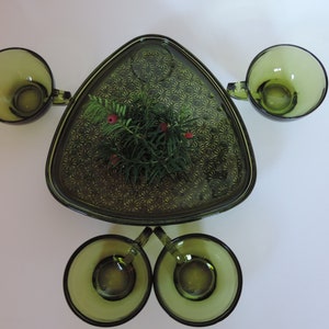 May include: A set of four olive green glass cups arranged around a central triangular glass serving tray. The tray features a repeating starburst pattern and holds a small sprig of evergreen needles with tiny red berries in the center.