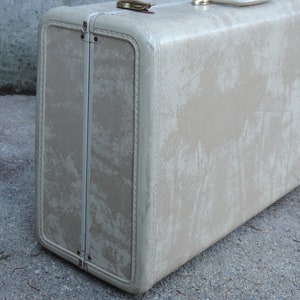 May include: Vintage beige suitcase with a rectangular shape and rounded corners. The suitcase has a distressed appearance with visible wear and tear. Features include a metal clasp and handle, suggesting it's a travel item. The exterior shows a faded, mottled pattern.