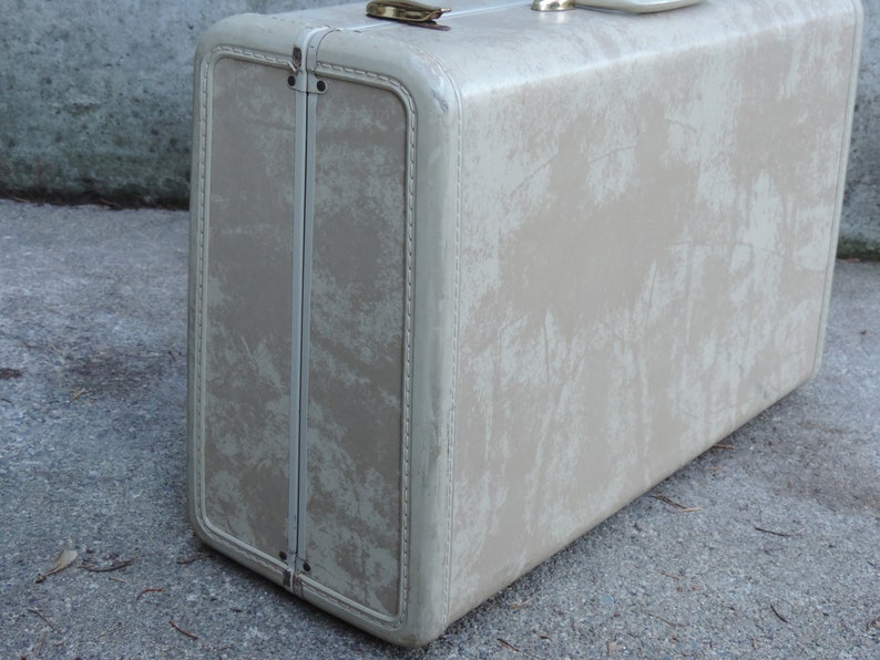 May include: Vintage beige suitcase with a rectangular shape and rounded corners. The suitcase has a distressed appearance with visible wear and tear. Features include a metal clasp and handle, suggesting it's a travel item. The exterior shows a faded, mottled pattern.