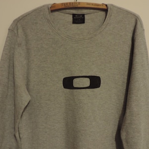 May include: A light gray long-sleeve sweatshirt with a black Oakley logo in the center. The sweatshirt is made of a ribbed material and has a crew neck. The Oakley logo is a black rectangle with rounded corners.
